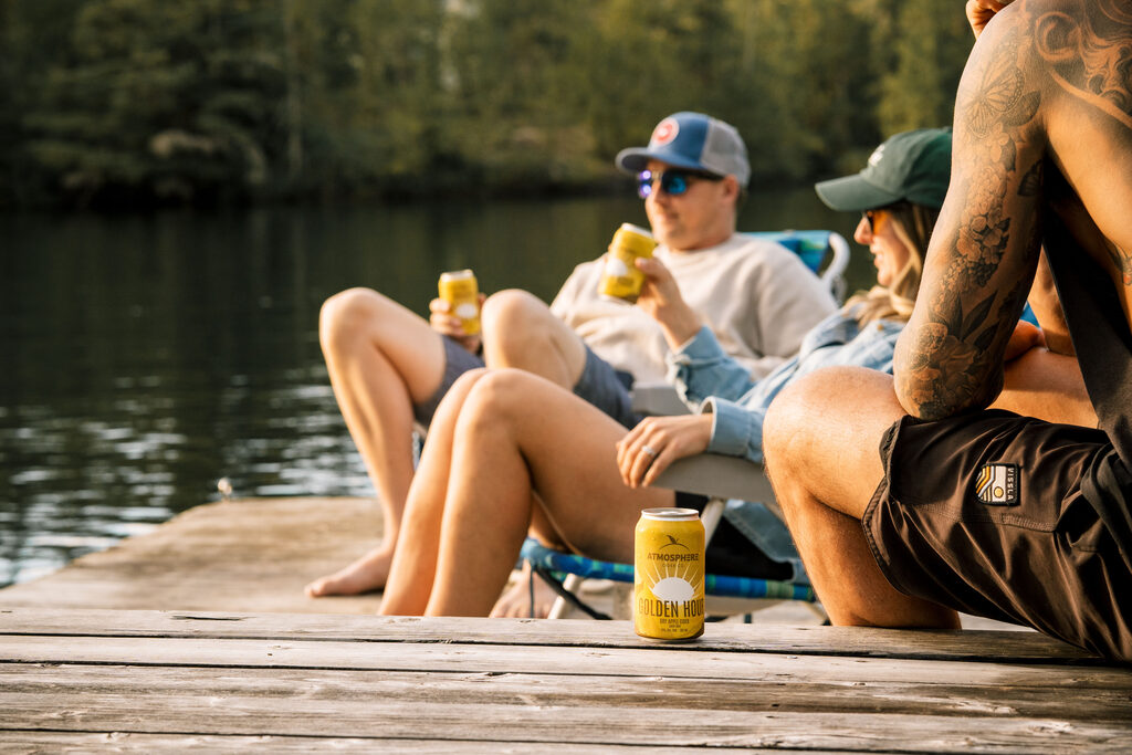 Friends relaxing in camp chairs on a lakeside dock with cans of Golden Hour